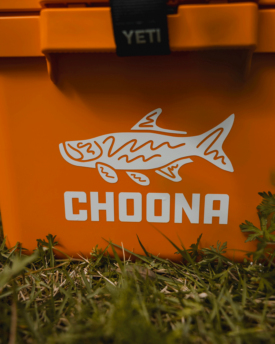 An orange Choona cooler with a white tarpon decal and CHOONA printed on it sits on grass, featuring the Choona Tarpon Decal. The YETI logo is visible on the top latch.