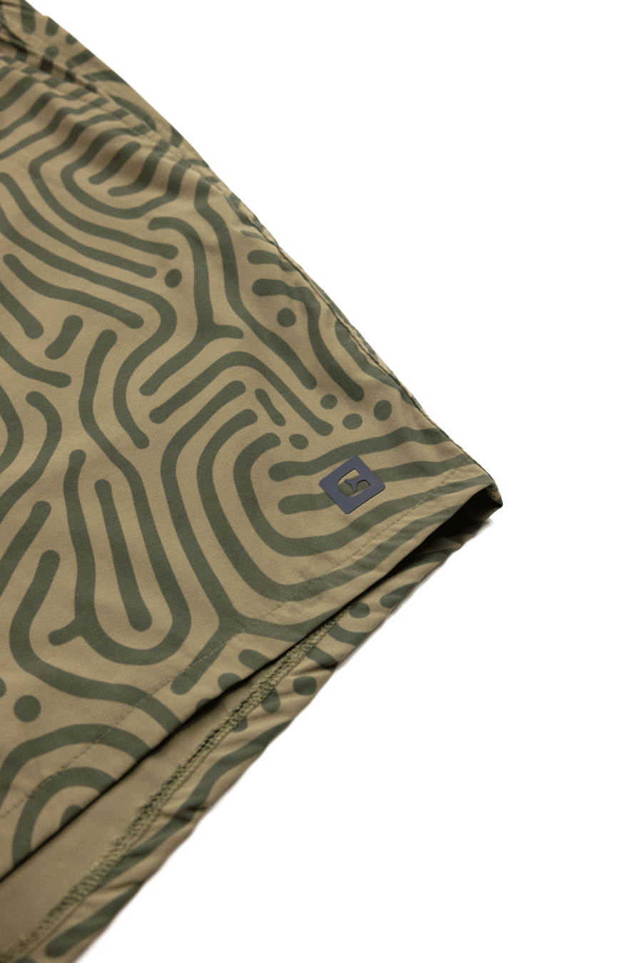 Close-up of Choonas Shore Short fabric, showing an abstract green and olive maze pattern. A small gray square logo with the letter S appears near the hem—perfect for coastal activities.