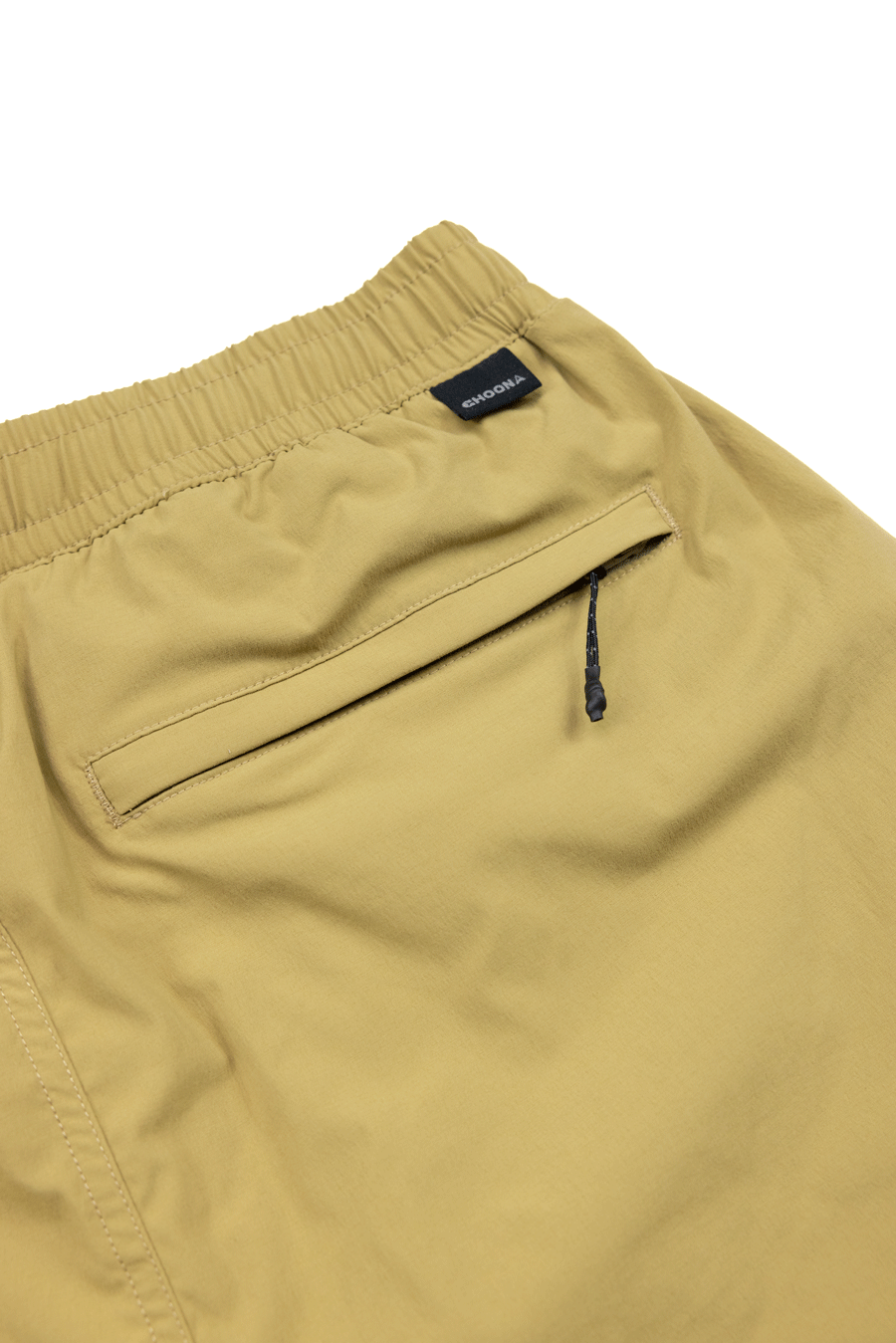 Close-up of the back of tan Choona Shore Short pants with an elastic waistband, zippered pocket with black pull, and a small black tag with white text—perfect for coastal activities.