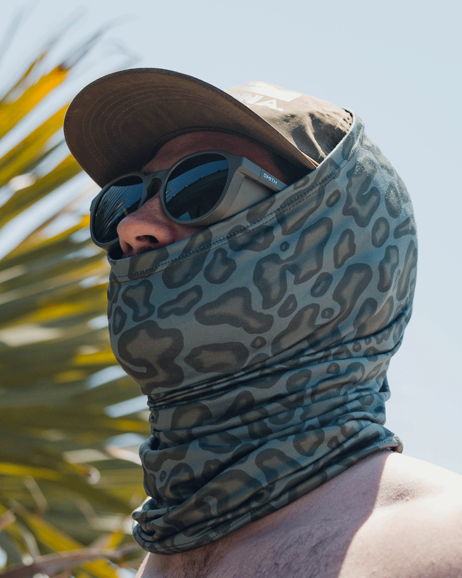 Wearing sunglasses, a cap, and the Choona Grayton Sun Mask with a camouflage pattern for outdoor protection, a person stands near palm leaves under a clear sky.