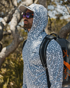 A man stands outdoors among trees, looking ahead, wearing the Choona Grayton Ultra-Lightweight Hoodie for breathable performance, sunglasses, a cap, and a backpack.