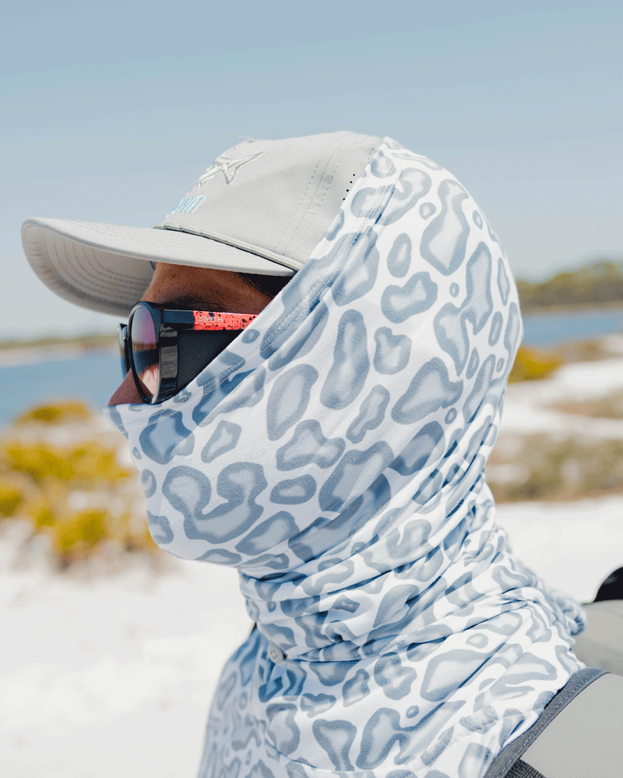 A person wears a Choona Grayton Sun Mask, paired with a light gray cap and dark sunglasses, while standing outdoors near a sunny beach with sand, water, and vegetation for enhanced outdoor protection.