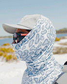 Wearing the ultra lightweight Choona Grayton Sun Mask, along with a gray cap and sunglasses, a person stands by water and sandy terrain on a sunny day, enjoying protection outdoors.