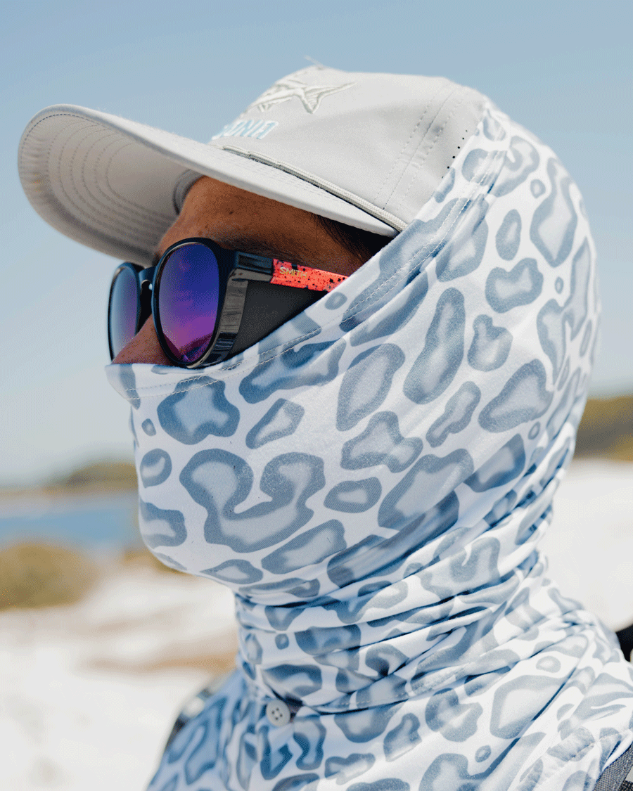A person wears a gray cap, reflective sunglasses, and the Choona Grayton Sun Mask—an ultra-lightweight animal-print mask—while standing outdoors on a sunny day with a blurred blue sky and landscape in the background.