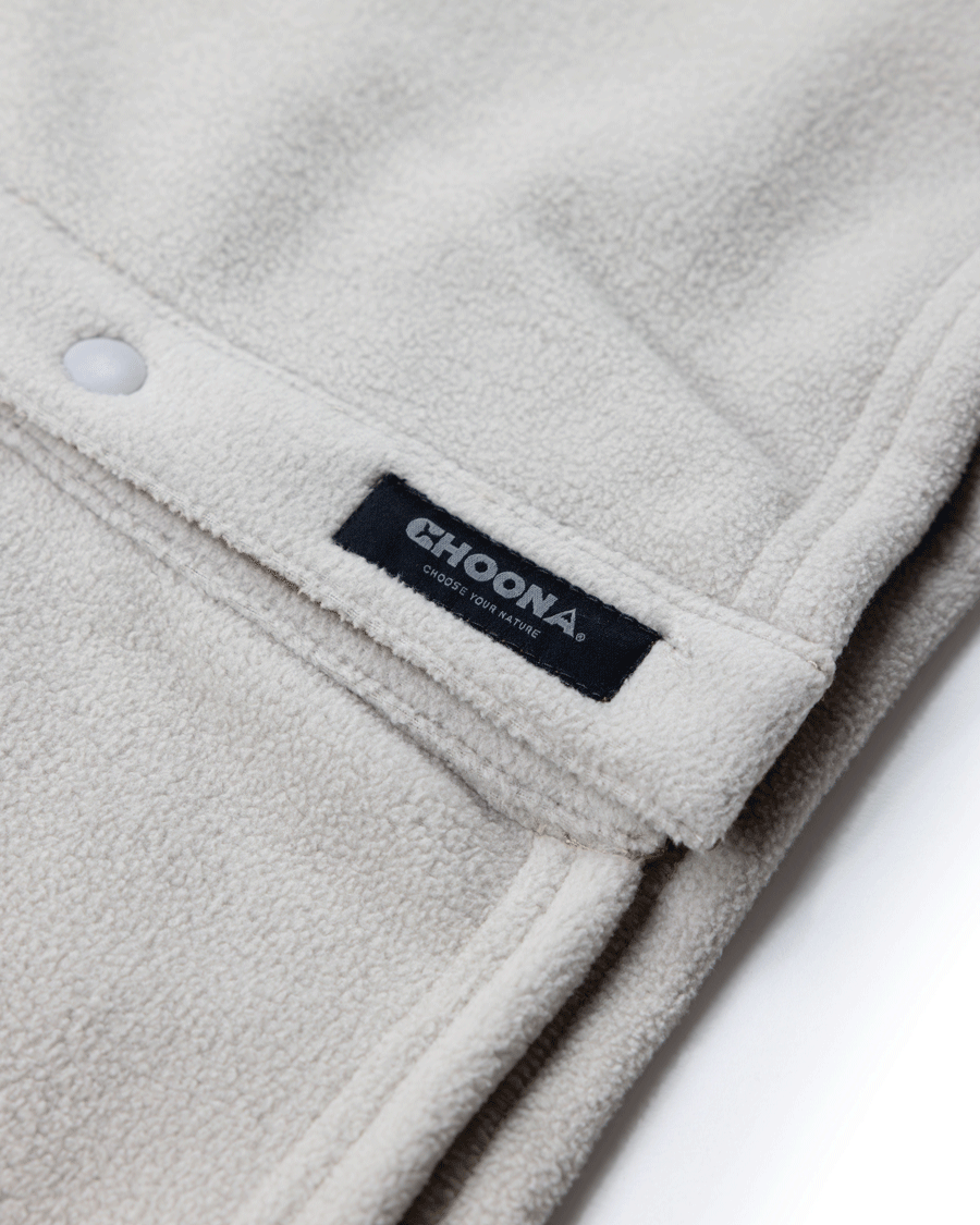 Close-up of the cream-colored Choona Skipper Shacket fleece, made from recycled polyester with a snap button and black label, shown on a white background—the perfect cold weather piece.