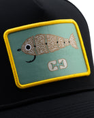 The Choona X Dent Redfish Snapback by Choona is a black hat with a rectangular yellow-edged patch featuring a tan and white cartoon fish on green, designed with fish bones and a white symbol below.