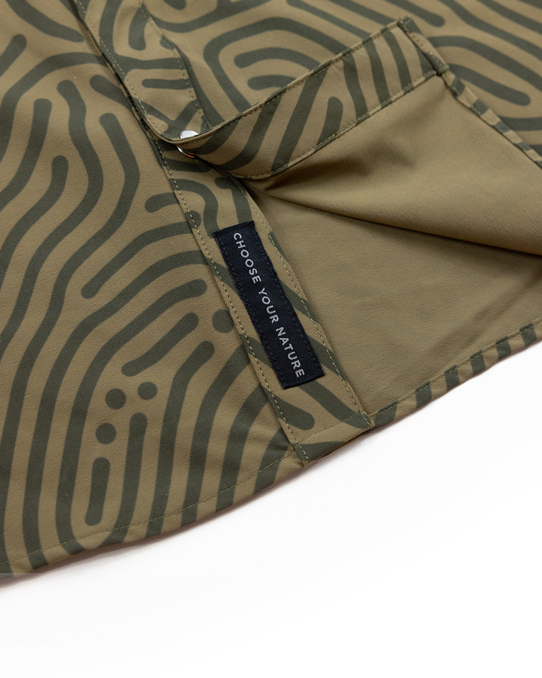 A close-up of olive-green fabric with black wavy patterns and a CHOOSE YOUR NATURE tag showcases the Choona Panhandle Pearl Snap, hinting at its signature pocket and inner lining for a unique coastal cowboy style.