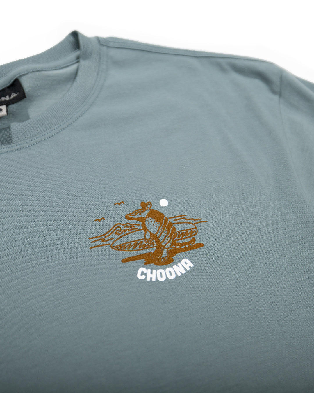 The Choona Armachillo Graphic Tee is a light blue retro surf shirt featuring a small surfing armadillo graphic with waves and sun, made from soft USA-grown cotton and "CHOONA" printed below the illustration.