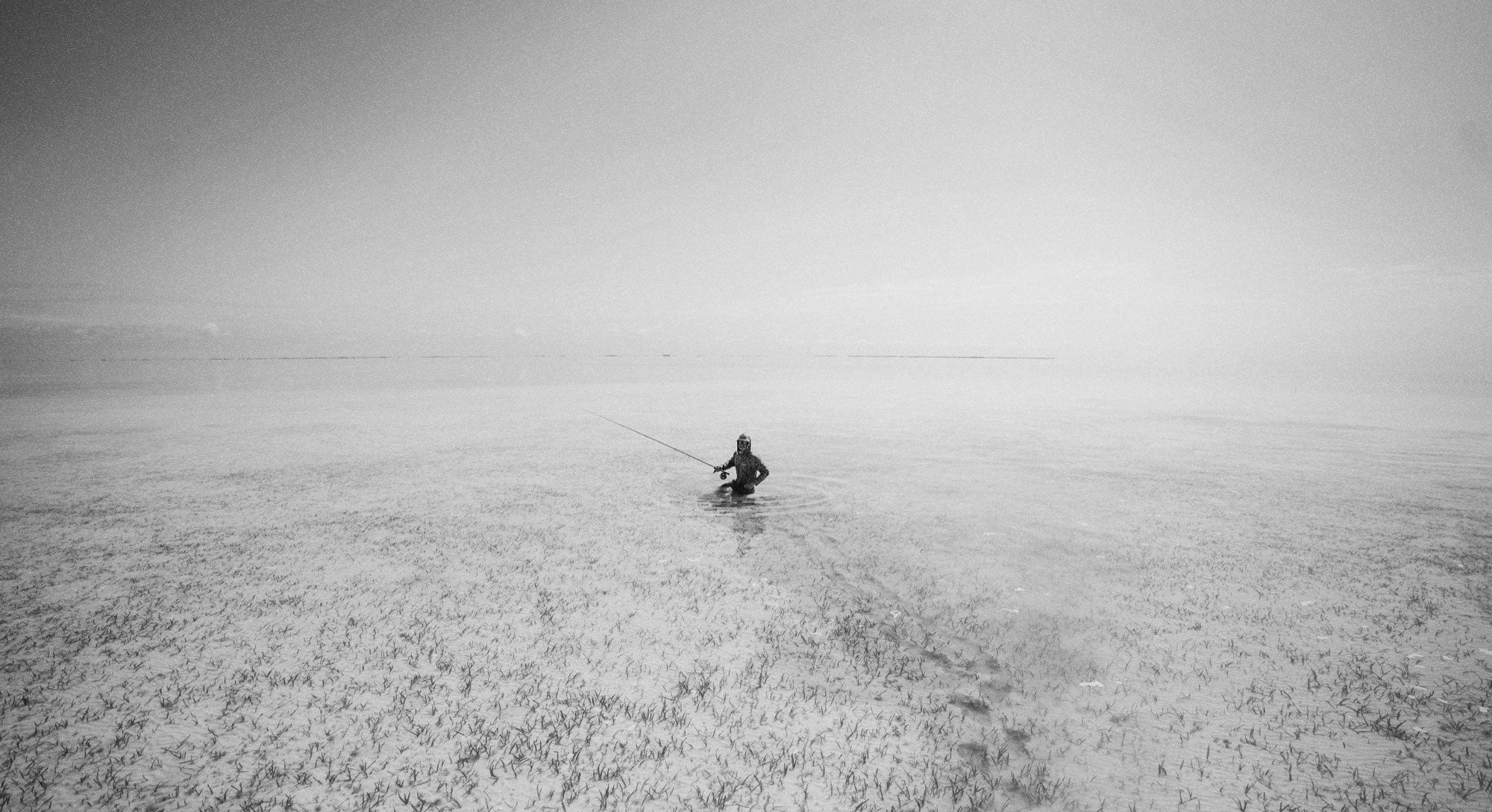 A person stands alone in shallow, clear water holding a fishing rod, surrounded by a vast, open landscape under a bright sky. The image is black and white, creating a serene, minimalist scene.