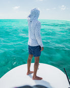 Wearing the Choona Grayton Ultra-Lightweight Hoodie and navy shorts, a person stands barefoot at the edge of a boat, gazing over vibrant turquoise ocean water beneath a bright sky.
