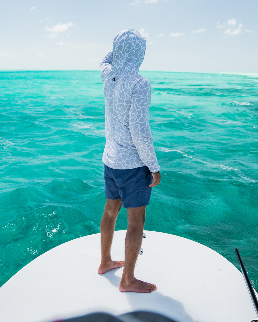 Wearing the Choona Grayton Ultra-Lightweight Hoodie and navy shorts, a person stands barefoot at the edge of a boat, gazing over vibrant turquoise ocean water beneath a bright sky.