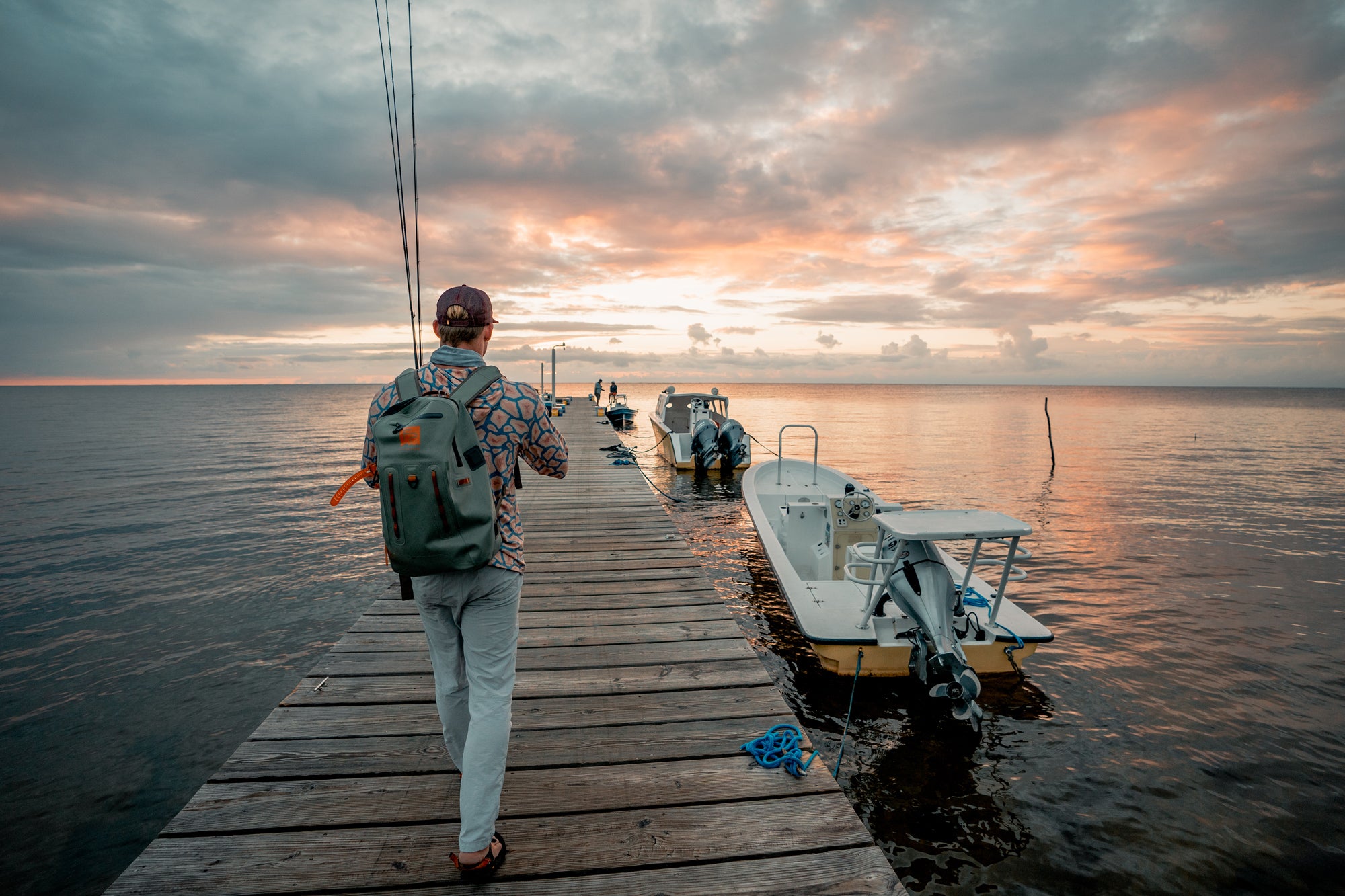 A person with a backpack walks along a wooden dock toward small boats at sunset, with calm water on both sides and a colorful sky filled with clouds.