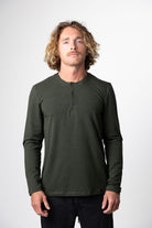 A man with wavy, shoulder-length blond hair wears Choona’s Versatile LS Henley in dark green and black pants, standing against a plain white background and looking directly at the camera with a neutral expression.