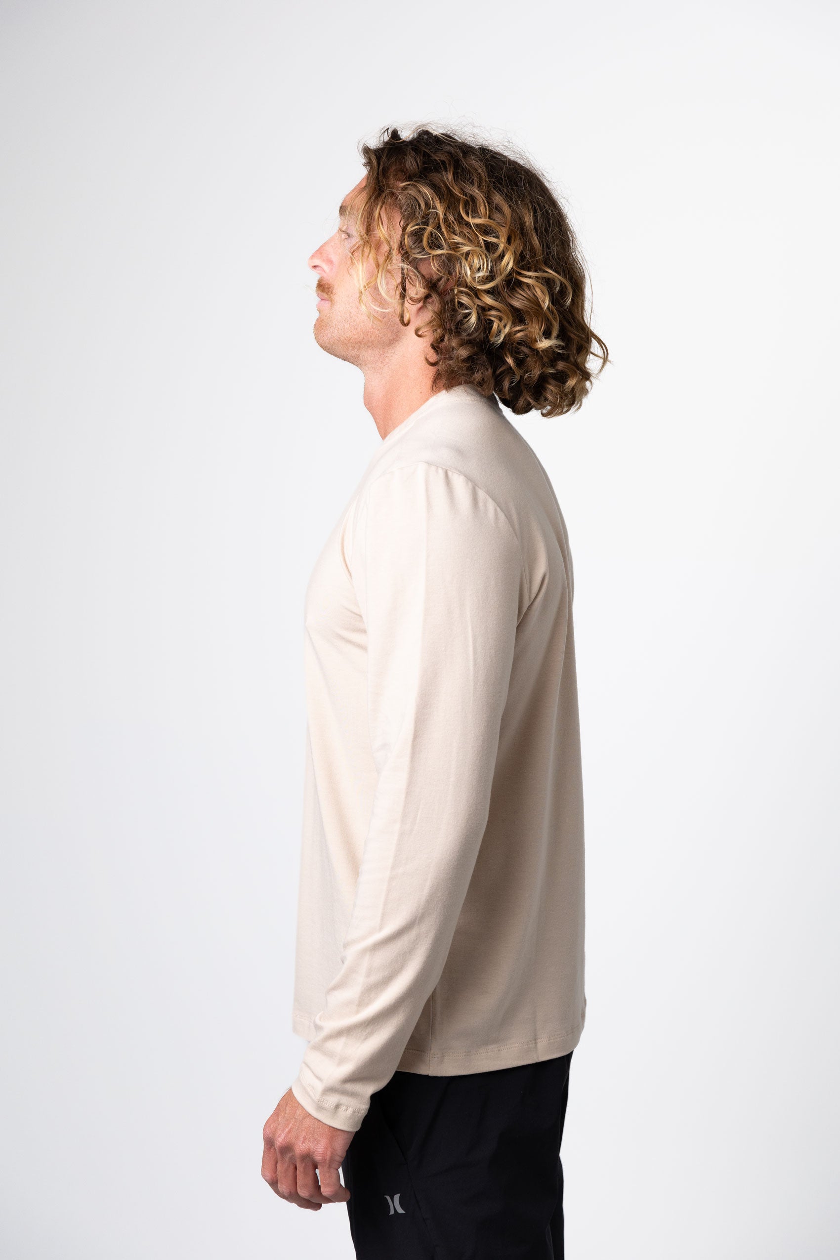A man with curly, shoulder-length hair stands sideways against a plain white background, wearing the Choona Versatile LS Henley in beige and black pants.