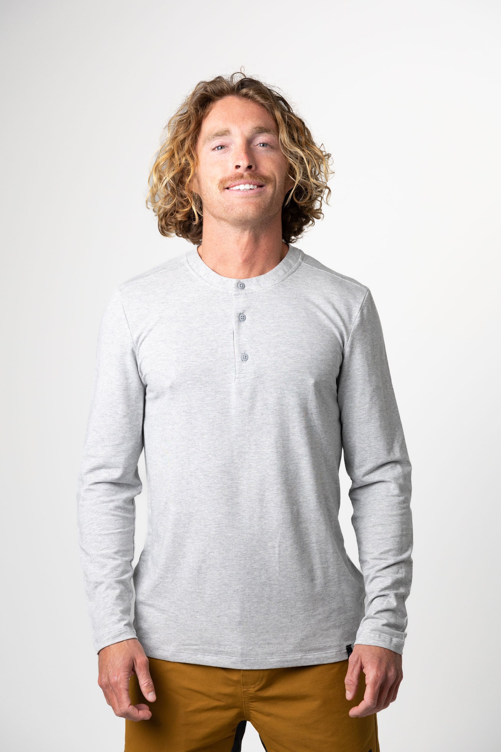 A man with curly, shoulder-length blonde hair smiles against a plain white background, wearing Choonas Versatile LS Henley in light gray—an outleisure staple—paired with mustard-colored pants.