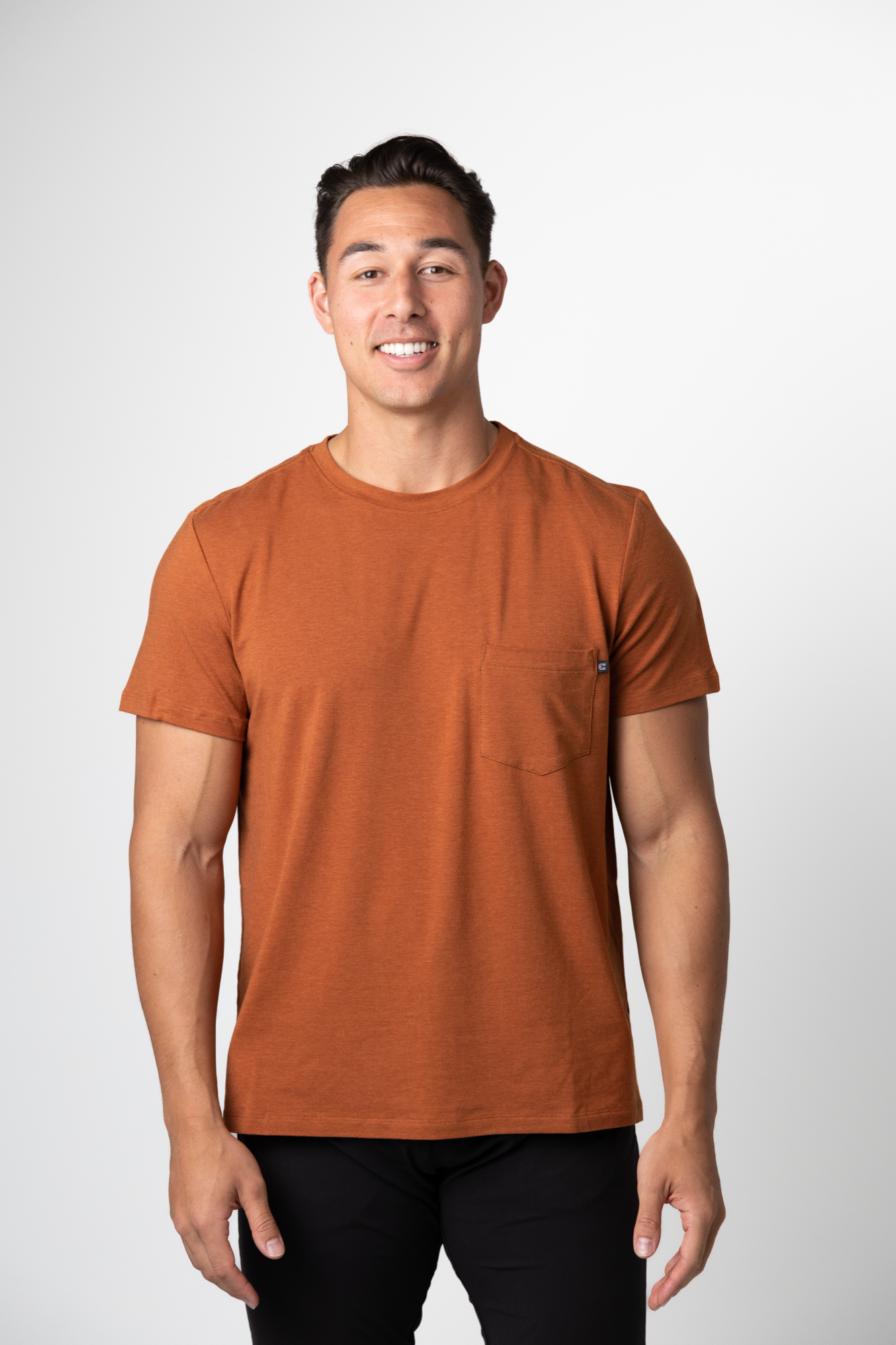 A man with short dark hair, smiling against a plain white background, is wearing a rust-colored Choona Panhandle Pocket Tee and black pants.