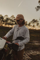 Wearing the Choona Versatile LS Henley, a person in a tan cap and sunglasses stands outdoors at sunset beside a white Suzuki motorcycle, embracing adventure among trees and clouds.
