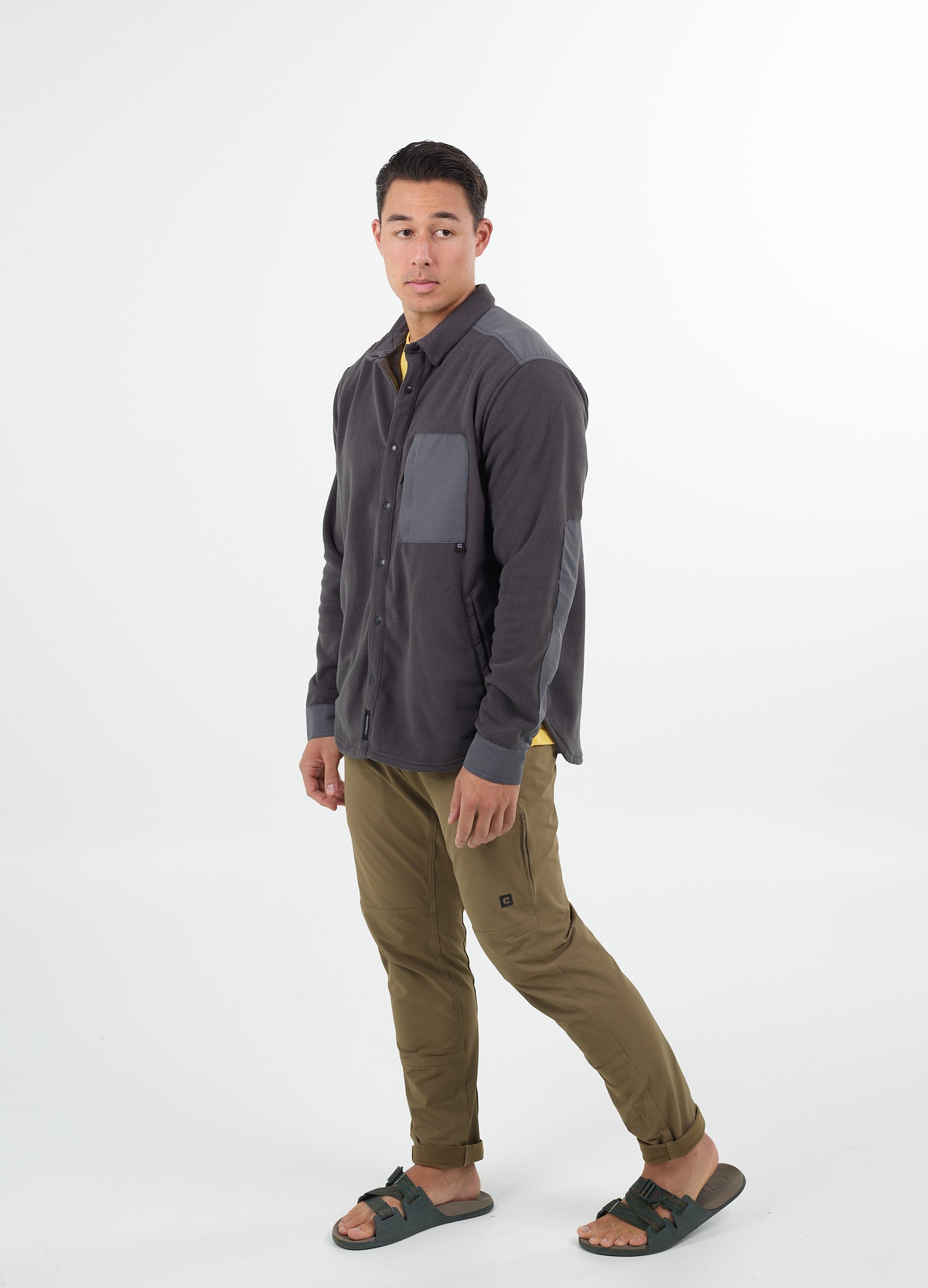 A man stands facing slightly left, wearing Choonas charcoal Skipper Shacket made from recycled polyester with a gray chest pocket, paired with khaki pants and green sandals. The background is plain white.